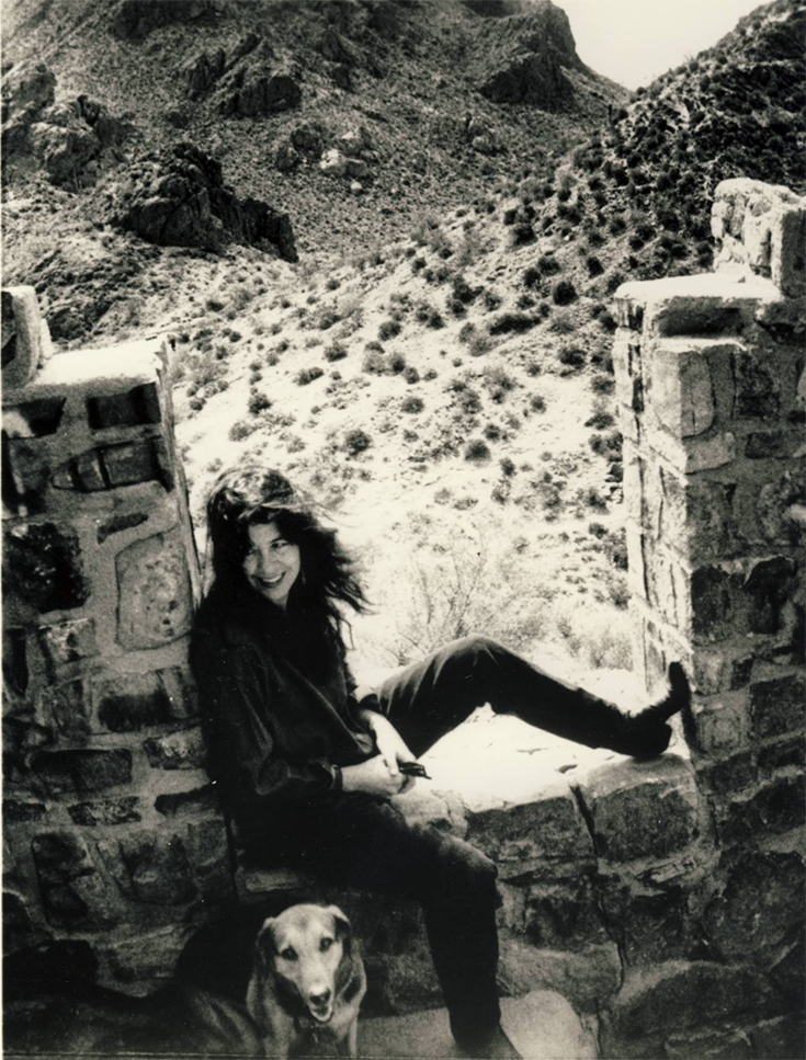 Poet Joy Harjo sits in the square frame of a brick wall in the desert, one leg on the sil. There is a dog sitting on the ground in front of her