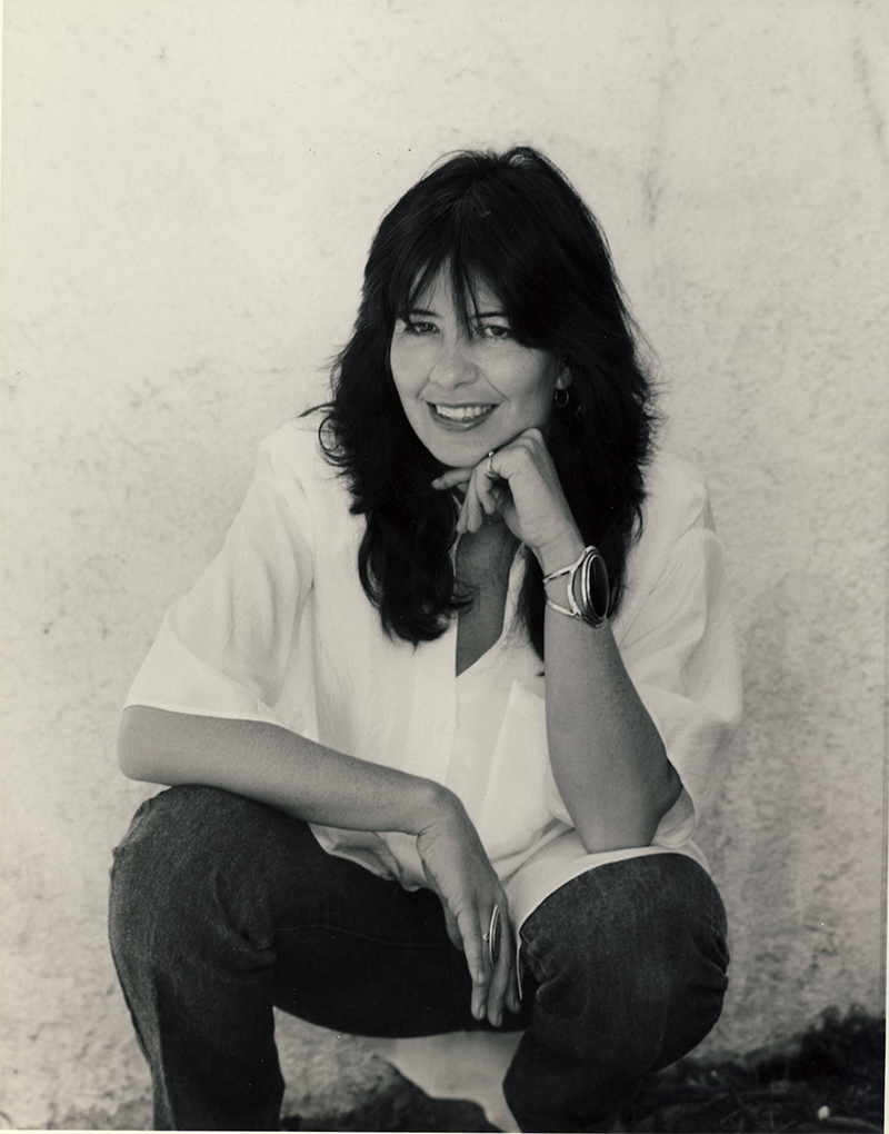 November 1989, University of Arizona, Tucson, AZ State University in Tempe. Poet Joy Harjo squats against a stucco wall wearing jeans and a white blouse, her head perched on her hand