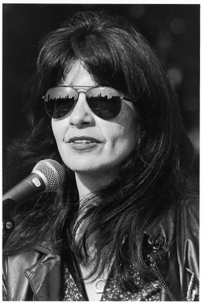 Poet Joy Harjo in front of a mic on stage wearing sunglasses with a city skyline reflected in them