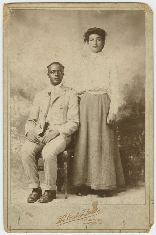 Studio portrait of a young couple, he seated, she with hand on his shoulder.